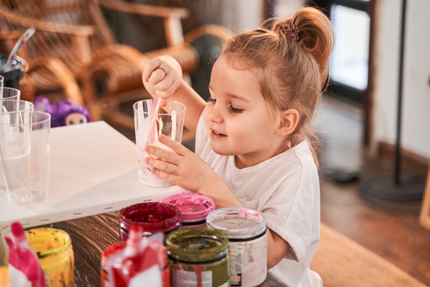 Dívka stojí u stolu a drží sklo s barvami, zatímco kreslení na umělecké škole - Fotografie, Obrázek