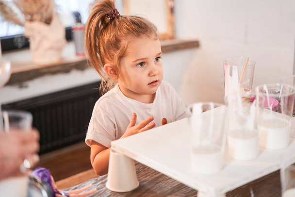 Female child looking at somebody attentively while standing at the table with paints - Foto, Bild