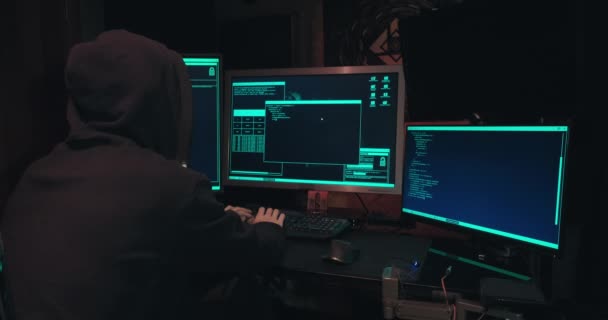 Hacker en una capucha se sienta frente a las pantallas de la computadora y hacks bases de datos - Metraje, vídeo