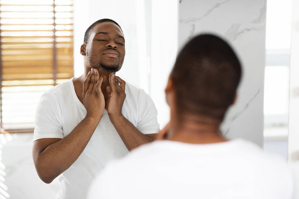 Gezondheidszorg Concept. Bezorgde zwarte man aanraken van de nek, onderzoeken lymfeklieren in de buurt van spiegel - Foto, afbeelding