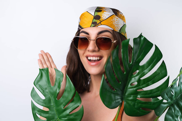 Sommerporträt einer jungen Frau im Sportbadeanzug, Kopftuch und Sonnenbrille mit Monstera-Palmblatt - Foto, Bild