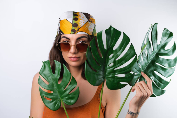 Sommerporträt einer jungen Frau im Sportbadeanzug, Kopftuch und Sonnenbrille mit Monstera-Palmblatt - Foto, Bild