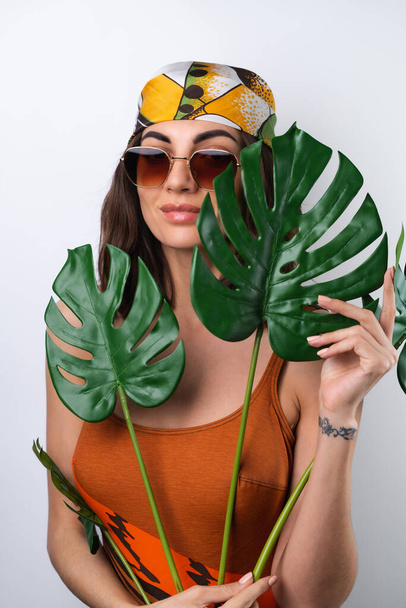 Summer portrait of a young woman in a sports swimsuit, headscarf and sunglasses with monstera palm leaf - Fotografie, Obrázek