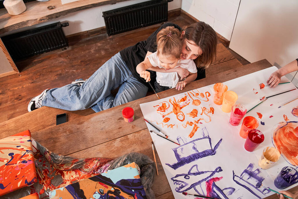 Mujer abrazándose con niña mientras dibuja junto con pinturas acrílicas en el papel - Foto, Imagen