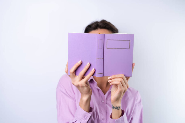 A young woman in a lilac shirt on a white background holds a book in her hands in glasses for vision - Foto, afbeelding