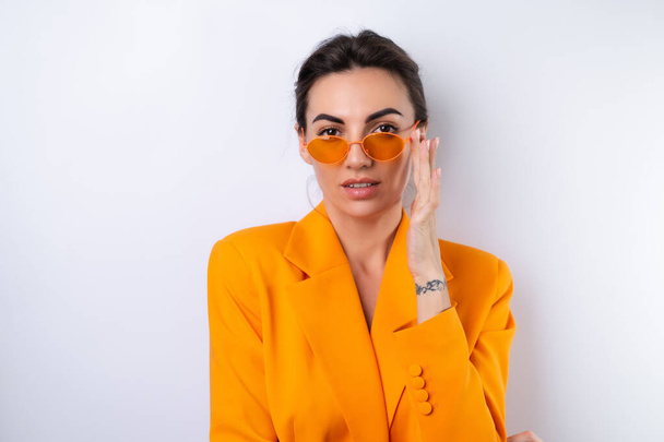Молодая женщина в модных стильных очках и ярко-оранжевой куртке негабаритного размера на белом фоне - Фото, изображение