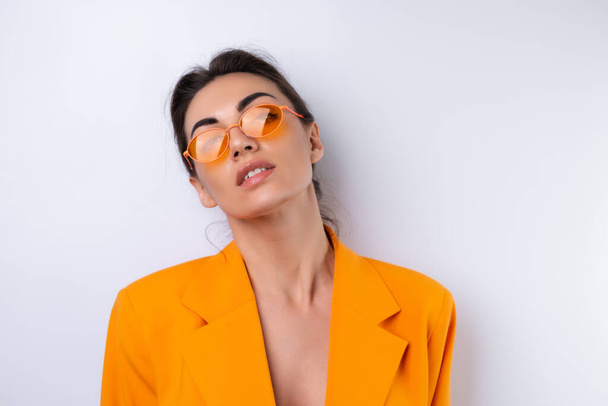 Jovem mulher na moda óculos elegantes e uma jaqueta laranja brilhante de grandes dimensões em um fundo branco - Foto, Imagem