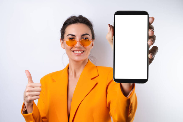 A young woman in trendy stylish glasses and a bright orange oversized jacket on a white background holds a phone with a blank white screen - Фото, изображение