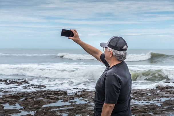 Turista adulto maduro con el pelo gris y una taza tomando un selfie en las rocas con el mar detrás y un cielo con nubes. - Foto, imagen