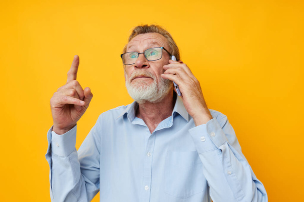 Portrait of happy senior man in a blue shirt and glasses talking on the phone yellow background - Photo, Image