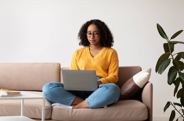Kanepede bacak bacak üstüne atmış vaziyette oturan odaklanmış genç siyahi bir bayan evde online iş veya eğitim için dizüstü bilgisayar kullanıyor. - Fotoğraf, Görsel