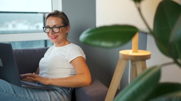 Smiling woman with glasses is sitting on the couch and working on a laptop or chatting with someone. Concept of remote work. - Footage, Video