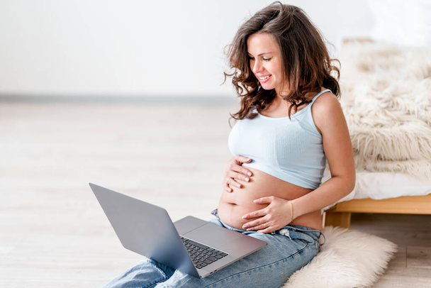 Une femme caucasienne enceinte est assise près du lit et travaille à distance à partir de la maison en utilisant un ordinateur portable ou en magasinant en ligne pour un bébé. - Photo, image