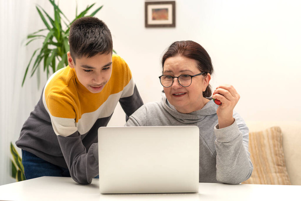Shot of schoolboy wearing yellow shirt helping his grandmother with glasses deal with the laptop  indoor. Grandmother smiles and holds an antistress ball in her hand from excitement - Fotografie, Obrázek
