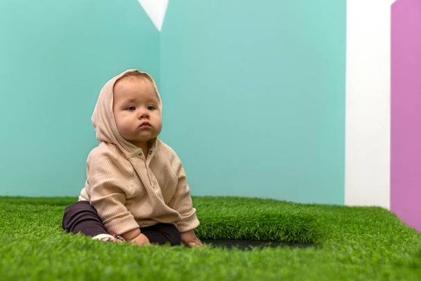 Full length portrait of serious baby wearing beige sweater with a hood sitting on artificial green grass against the background of colored walls inside - Foto, afbeelding