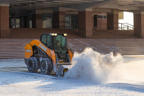 Krasnojarsk, Russland - 15. Februar 2022: Kleines Schneeräumfahrzeug auf dem Stadtplatz. Gelber Traktor beim Schneeräumen auf einer Straße. Lademaschine beseitigt Schnee im Winter - Foto, Bild
