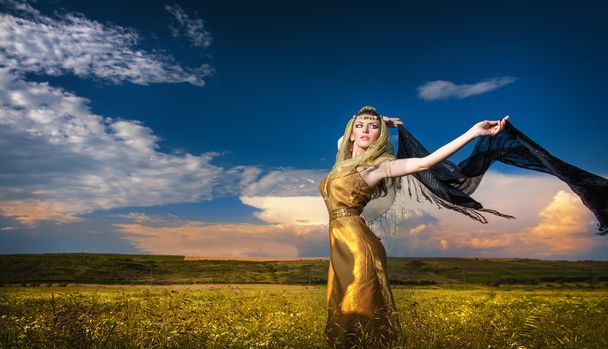 Lovely young lady posing dramatically with long black veil on green field. Blonde woman with cloudy sky in background - outdoor shot. Glamorous female in nature - Arabian style - Photo, image