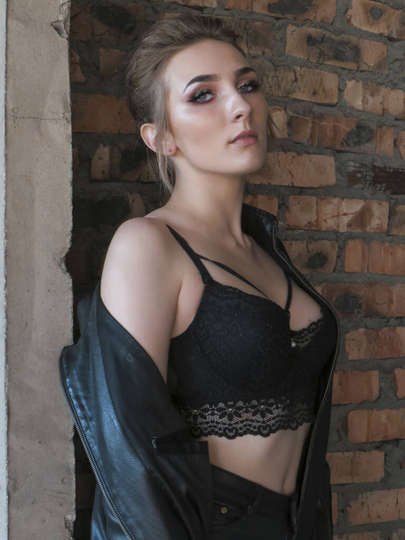 A young attractive woman with a slim body in a black leather jacket and leggings and a black bra top against a brick wall in an abandoned building - Photo, Image