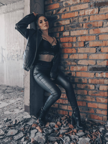 A young attractive woman with a slim body in a black leather jacket and leggings and a black bra top against a brick wall in an abandoned building - Foto, Imagen