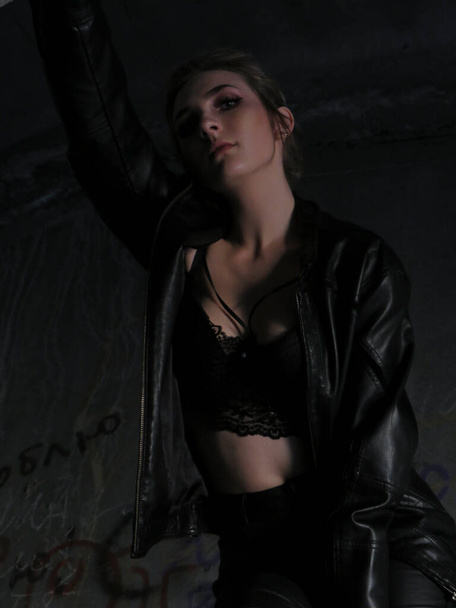 A young attractive woman with a slim body in a black leather jacket and leggings and a black bra top against a brick wall in an abandoned building - Foto, Imagem