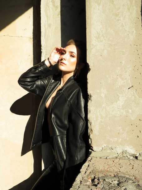 Attractive young woman with a slim athletic body in a black leather jacket and leggings at the window opening in the sunlight inside a non-residential building, art photo, beauty and health concept - Photo, Image