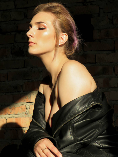 Attractive young woman with a slim athletic body in a black leather jacket and leggings against a brick wall in the sunlight inside a non-residential building, art photo, catwoman concept, beauty and health concept, body care - Fotografie, Obrázek