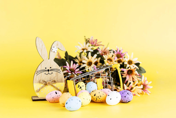 Színes húsvéti fürj tojás bevásárlókocsiban, fa nyuszi és tavaszi krizantém virágok sárga alapon. Másold a helyet. Minimális húsvéti fogalom. - Fotó, kép