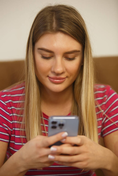 Young blonde woman texting online in messenger app on modern mobile phone. Cheerful white female with long blond hair typing message on social media application in blue smartphone with triple camera - Zdjęcie, obraz