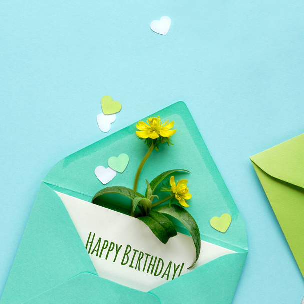 Happy Birthday card. Spring background in shades of green. Paper envelope with card and yellow erranthis flowers, heart confetti on mint green paper. Square composition. - Foto, afbeelding