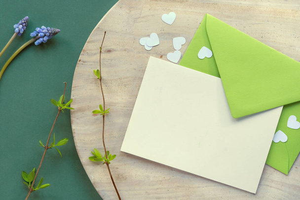 Spring background in shades of green. Paper envelope with blank greeting card and heart confetti on beige stone on dark green table. Fresh spring leaves on twigs. - Foto, immagini