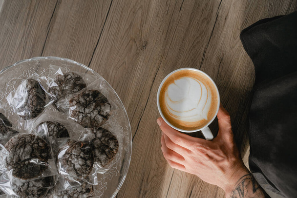 top view of coffee in a cup, chocolate biscuits lie nearby, a barista's hand with a tattoo holds a cafe cup - Photo, Image