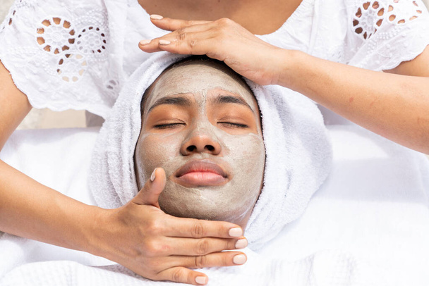 Afroamerykanka leżąca maseczkę z masażystką leczącą twarz w salonie spa przy basenie. - Zdjęcie, obraz