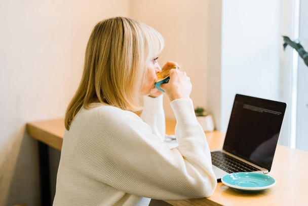 Зрелая взрослая женщина, сидящая в кафе, пьющая кофейную кружку и работающая онлайн на ноутбуке. Бизнесмен в очках, печатающая на ноутбуке, в коворкинге в кофейне - Фото, изображение