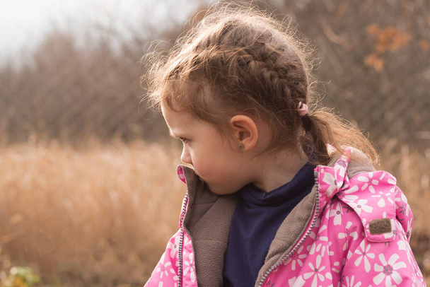 Ένα μικρό όμορφο κορίτσι σε ένα σακάκι με ξανθά μαλλιά σε κοτσίδες στέκεται σε ένα φθινοπωρινό φόντο δυστυχώς κοιτάζοντας κάτω - Φωτογραφία, εικόνα
