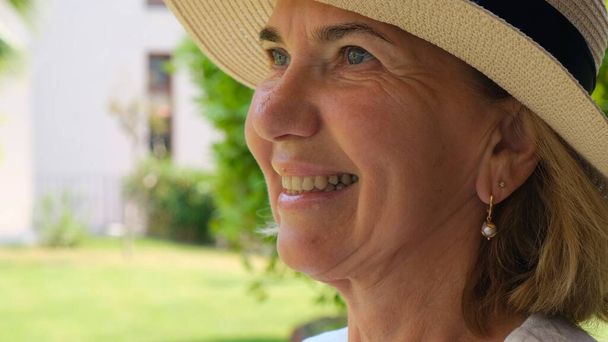 Porträt einer reifen Frau zwischen 55 und 60 Jahren mit einem zahmen Lächeln im sonnengeschützten Strohhut, die ihre Zeit vor der Kulisse eines grünen Gartens genießt. Nahaufnahme. - Foto, Bild