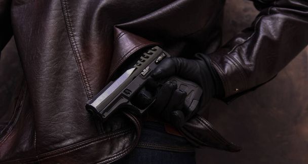 a man in a leather jacket and jeans holds a gun in his hand in a leather glove - Foto, Bild