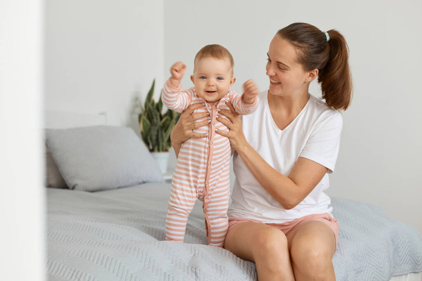 Portrait of positive optimistic young mother wearing white casual t shirt sitting on bed in bedroom with toddler baby, woman holding kid's hands, child standing near mom with happy expression. - Фото, зображення