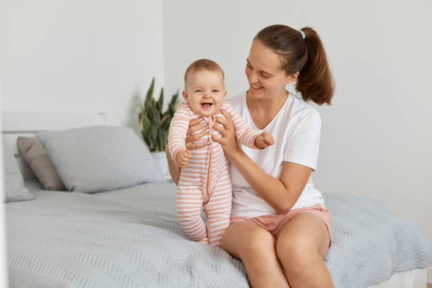 Indoor shot of dark haired woman wearing white casual t shirt sitting on bed in bedroom with toddler baby, mommy playing with child, looking at kid with love and gentle. - Zdjęcie, obraz