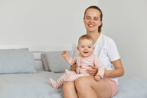Πορτρέτο της θετικής ενθουσιασμένοι νεαρή ενήλικη μητέρα φορώντας λευκό casual t shirt κάθεται στο κρεβάτι στο υπνοδωμάτιο με το μωρό, θηλυκό κρατώντας το παιδί της και κοιτάζοντας κάμερα με αισιόδοξα συναισθήματα. - Φωτογραφία, εικόνα