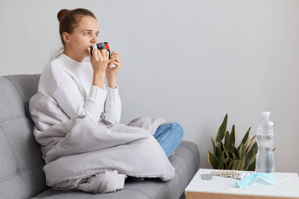 Indoor shot of unhealthy female wearing white sweater and bun hairstyle, sitting on sofa wrapped in blanket and drinking hot tea, having headache and high temperature. - Foto, Bild