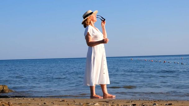 vrouw in een strohoed en witte jurk loopt langs de kust zet op zonnebril en bewondert de blauwe zee op een zonnige zomerdag, genieten van vrijheid en ontspanning. Het concept van een bejaarde burger - Foto, afbeelding
