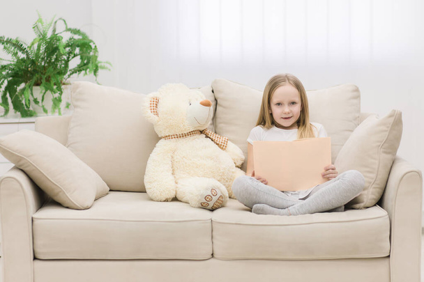Photo of plush teddy bear and little girl holding a book on sofa. - Fotó, kép