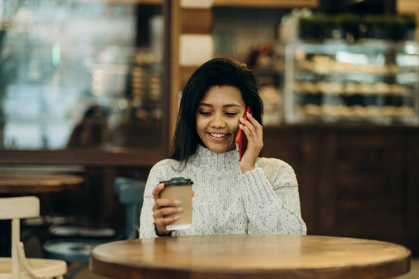 Афроамериканка в кафе разговаривает по телефону и пьет кофе. Темнокожая молодая женщина сидит в кафе и пьет кофе, ожидая своего партнера. - Фото, изображение