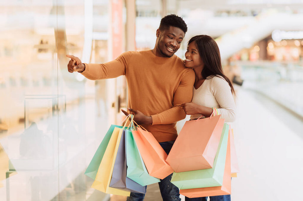 Die Wahl des passenden Geschenks. Lächelndes afroamerikanisches Paar, das in einem Einkaufszentrum spaziert, Schaufenster anschaut und mit dem Finger auf das Schaufenster zeigt. Saisonale Weihnachtsverkäufe, Rabatte und Sonderangebote - Foto, Bild