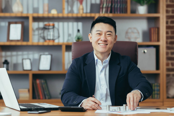 Portrait of a successful Asian banker, businessman working in the office, looking at the camera and smiling, counting metal coins on the table - Фото, изображение