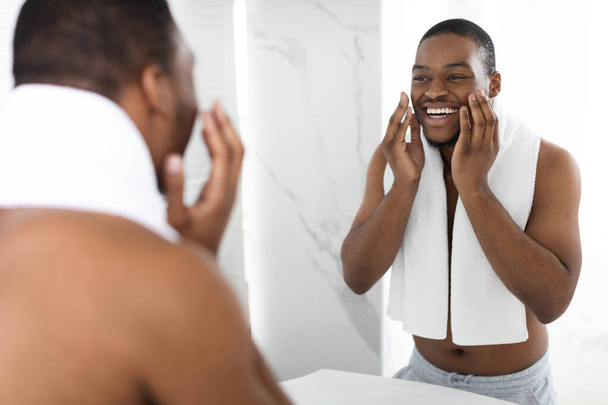 Tıraş losyonu. Banyoda hazırlanan mutlu gömleksiz zenci portresi, boynunda havluyla yakışıklı Afrikalı Amerikalı bir adam, evde günlük temizlik yapıyor, seçici bir odak noktası var. - Fotoğraf, Görsel