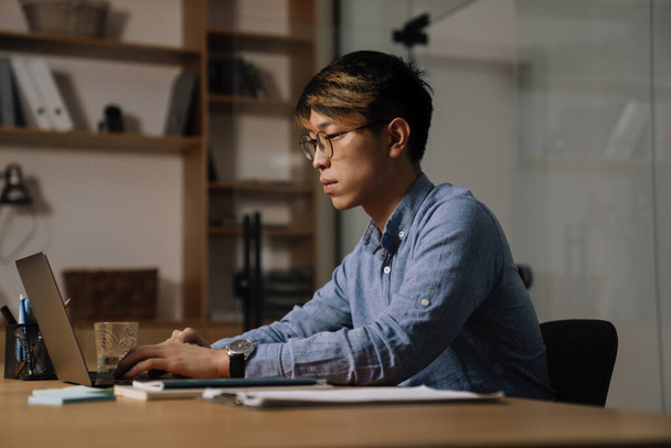 Focused asian guy in eyeglasses working with laptop while sitting at table in office - Fotoğraf, Görsel