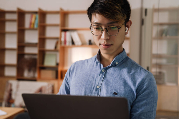 Focused asian guy in earphones working with laptop in office - Foto, Imagem