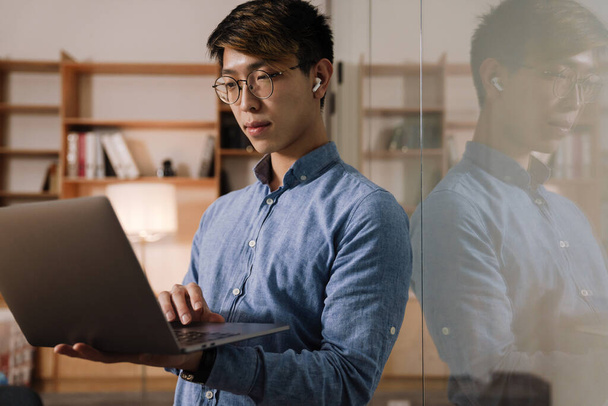 Focused asian guy in earphones working with laptop while standing in office - Foto, Imagen