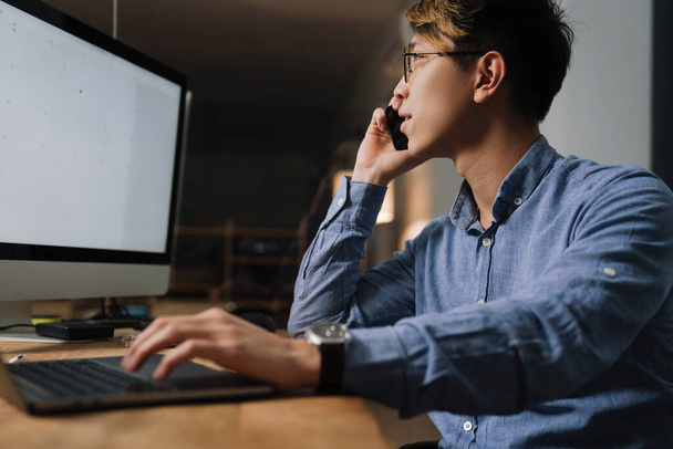Сфокусированный азиатский парень разговаривает по мобильному телефону во время работы с компьютерами в офисе - Фото, изображение